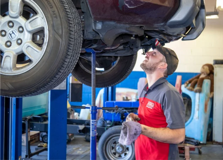 A Grease Monkey mechanic in uniform inspects the underside of a raised car in an auto repair shop, holding a rag and looking upward.