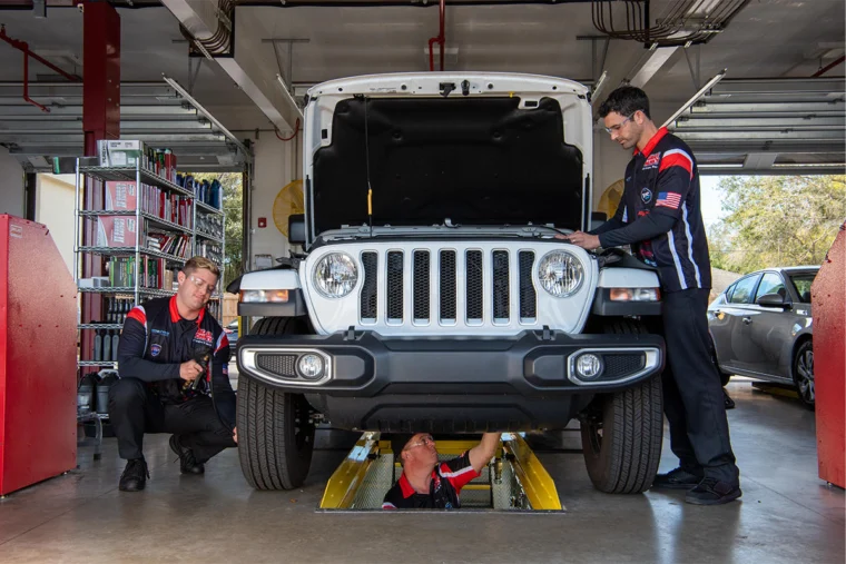 Three mechanics inspect a white Jeep in an auto repair shop; one is under the vehicle, while the other two stand and crouch beside it.