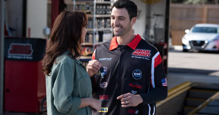 A man in a racing-themed Grease Monkey shirt talks with a woman in a green blouse in an automotive garage setting.