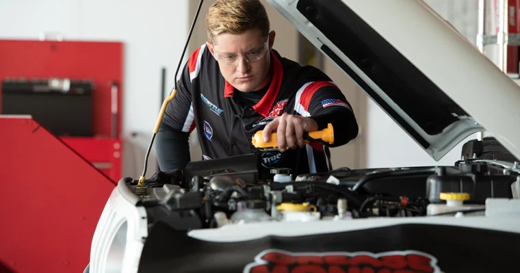 A person in safety glasses inspecting the engine under the hood of a white car in a garage or workshop.