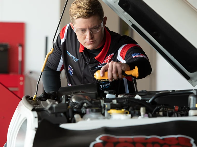 A person in safety glasses inspecting the engine under the hood of a white car in a garage or workshop.