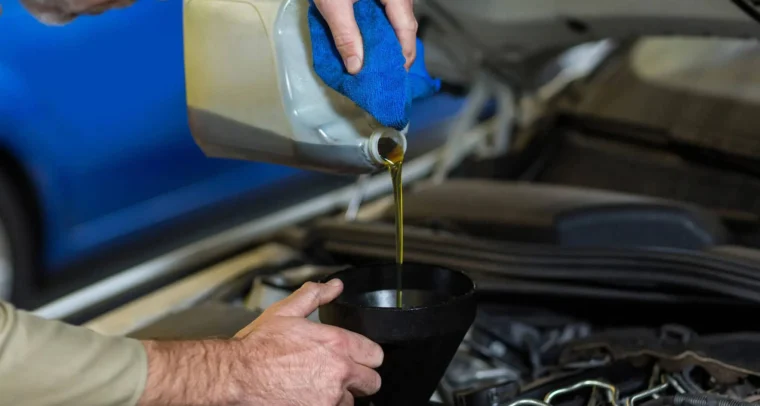 A person pours engine oil from a container into a black funnel placed in a car engine compartment.
