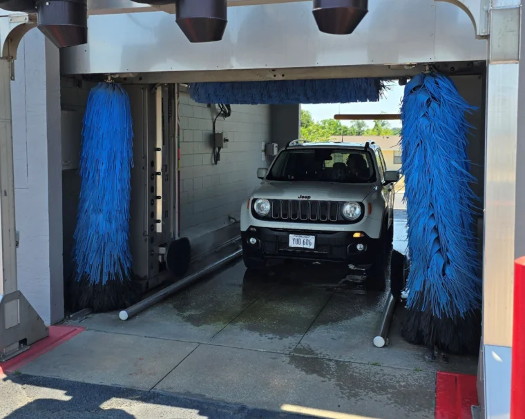 A white Jeep Renegade is positioned inside an automated car wash, surrounded by large blue cleaning brushes.