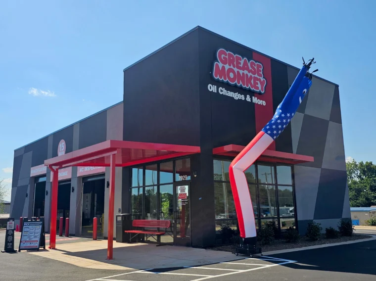 Exterior of a Grease Monkey service center with a red awning, a waiting bench, and a star-spangled inflatable tube man in front.