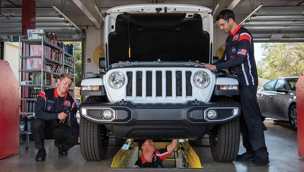 Three auto technicians inspect and work on a white SUV in a garage, with one technician beneath the vehicle and two others at the front and side.