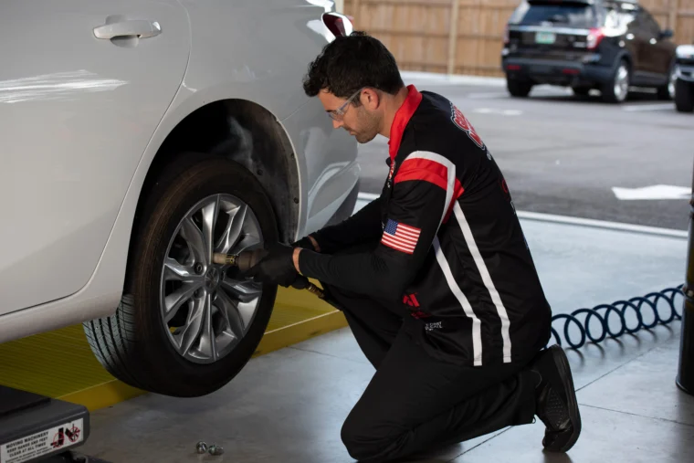 A mechanic kneels while using a power tool to remove a tire from a white car in an auto service garage.