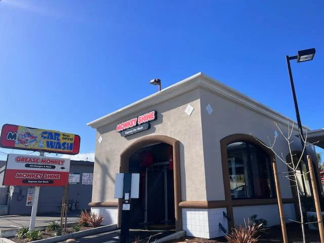 A small car wash building with a sign reading "Monkey Shine Car Wash" under a clear blue sky; a roadside sign advertises the business.
