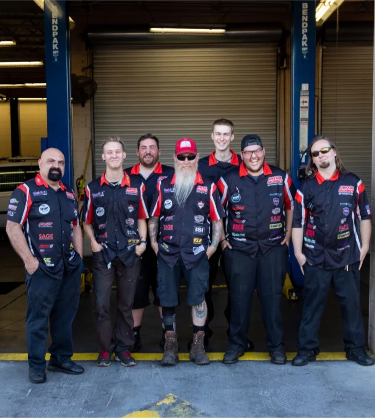 Six men in matching black and red team shirts stand together in a garage, posing for a group photo in front of a partially open roller door.