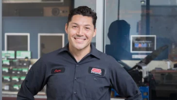 A man in a Grease Monkey uniform stands with hands on hips, smiling at the camera. A window shows equipment and shelves in the background.