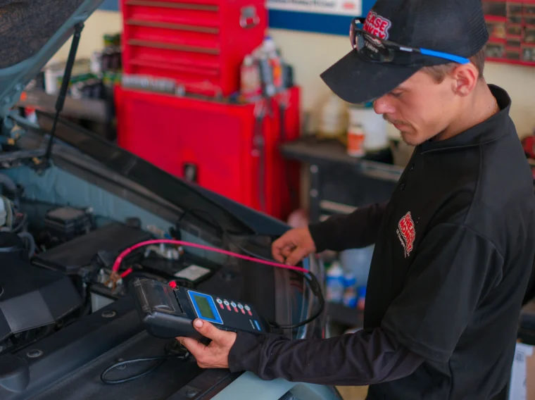 A mechanic uses an electronic diagnostic tool to check a car battery while performing Battery Services in a garage with a red tool cabinet in the background.