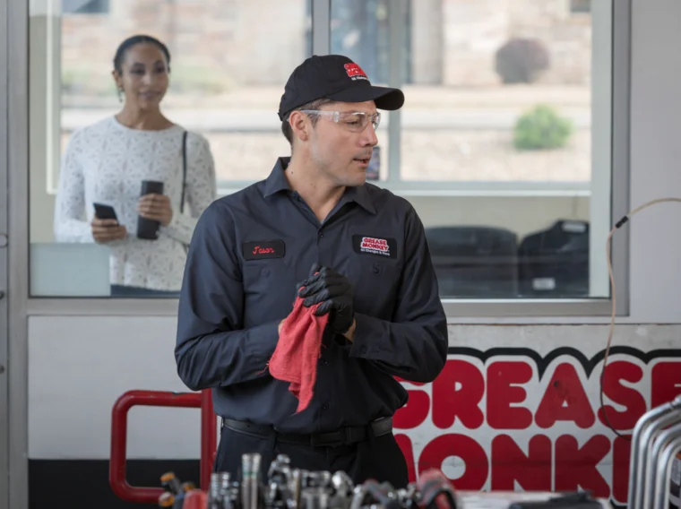 A mechanic wearing a uniform and gloves holds a red cloth in a garage, ready to provide Battery Services. A woman stands in the background holding her phone and a coffee cup, watching through a glass window.