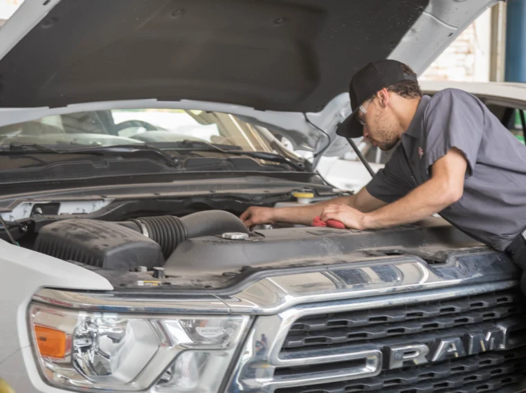 A mechanic wearing a hat and safety glasses inspects the engine of a white RAM truck with the hood open in a garage, performing routine checks and Battery Services.