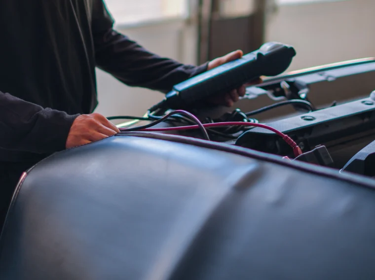 A mechanic wearing a cap uses a diagnostic tool under the hood of a car in a garage, checking essential components during battery services.