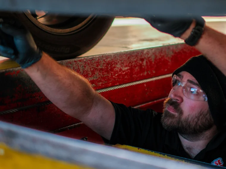 A man wearing safety glasses and gloves performs brake services on a vehicle tire, viewed through a gap between metal surfaces.