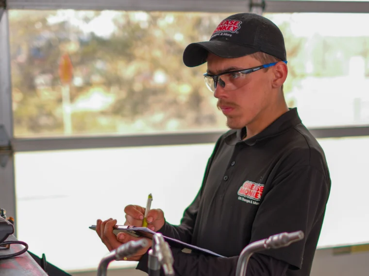 A man in a black Grease Monkey uniform and hat writes on a clipboard while standing near automotive equipment in a garage, preparing to perform expert brake services.