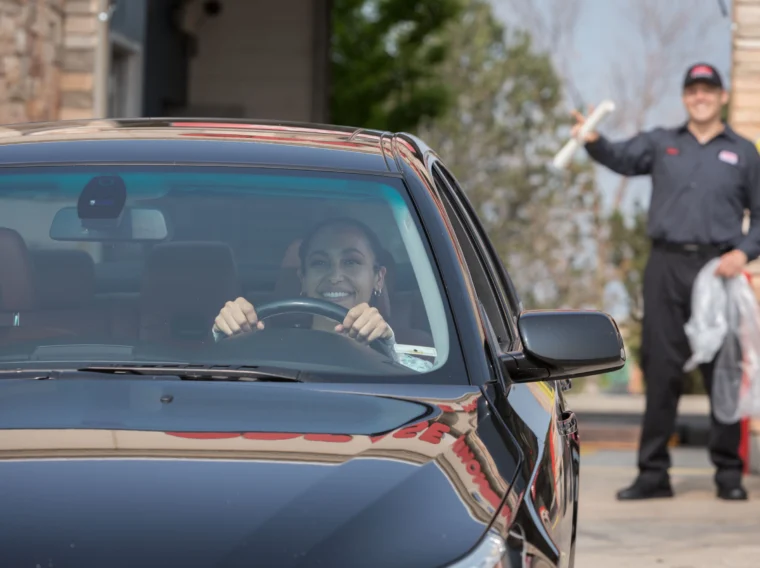 A woman smiles while sitting in the driver’s seat of a black car at a drive-thru Car Wash, as a uniformed worker stands outside holding a rolled-up document.