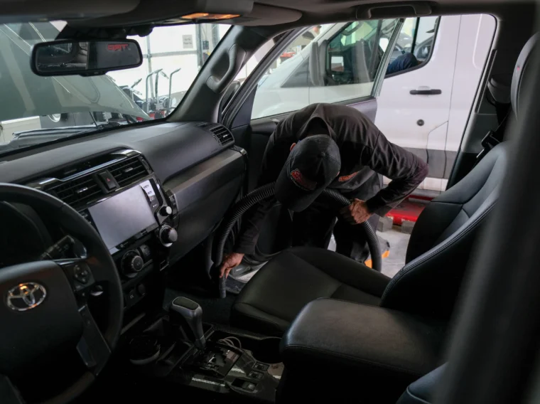 A person in a uniform is vacuuming the interior floor of a parked car at an auto detailing facility, showcasing professional car wash services.