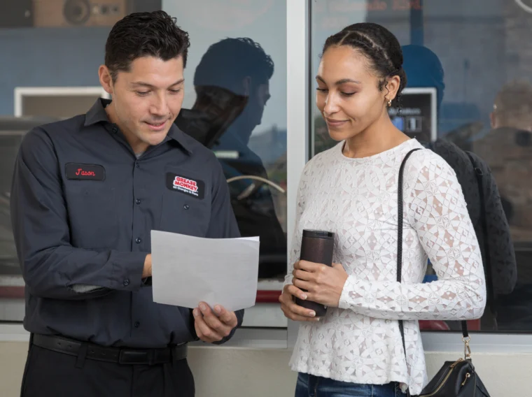 A man in a Grease Monkey uniform showing a document about clutch replacement to a woman holding a travel mug; they are standing near a window.