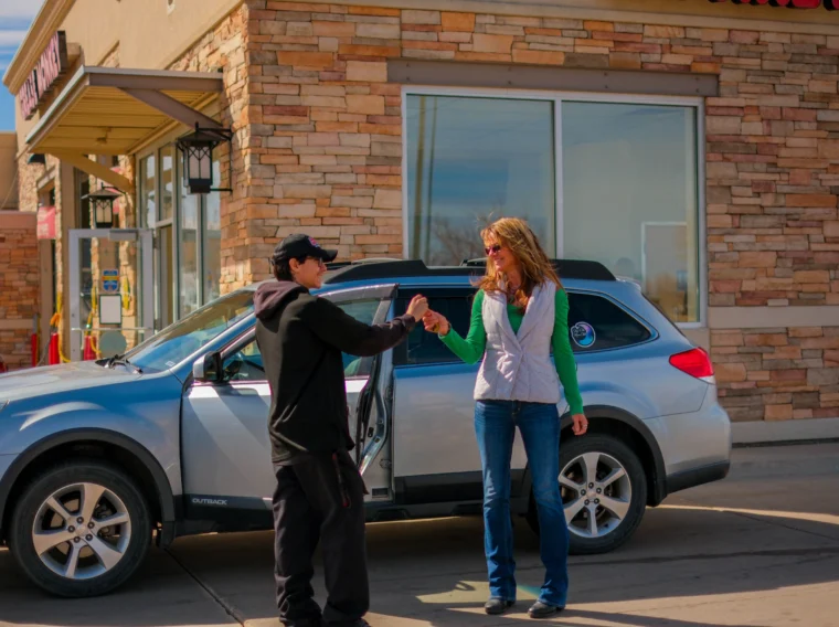 A woman and a man fist bump beside a silver car parked outside a Grease Monkey building with stone walls and large windows, celebrating a successful clutch replacement.