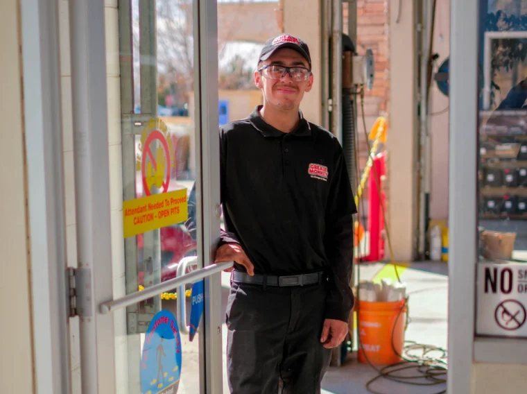 A man in a Grease Monkey uniform and hat stands at an open glass door, smiling, with a service area visible in the background—highlighting expert Engine Services offered on site.