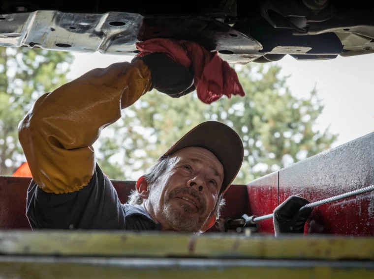 A mechanic wearing gloves and a cap wipes the underside of an EV with a red cloth while performing EV maintenance in a garage.