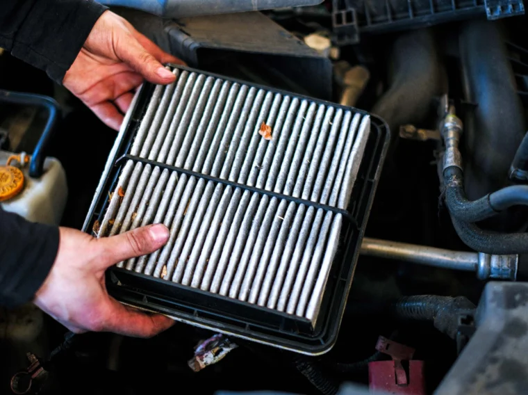 A person holds a rectangular car air filter above an engine bay, showing some dirt and debris trapped in the filter—a clear sign it's time to consider professional Filter Services.