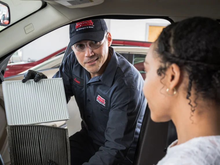 A Grease Monkey mechanic in uniform explains Filter Services to a woman inside a car, showing her two cabin air filters—one clean and one dirty—to illustrate the difference.