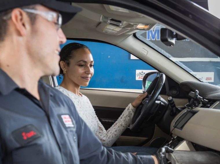 A Grease Monkey mechanic in uniform checks the glove compartment of a car as part of filter services while a woman sits in the driver’s seat, smiling.