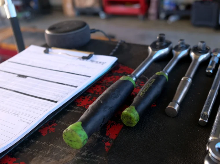 A clipboard with paperwork sits next to various ratchets and sockets on a workbench in a garage setting.