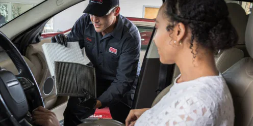 A mechanic shows a car's cabin air filter comparison to a seated customer, displaying a clean filter and a dirty one inside the vehicle.