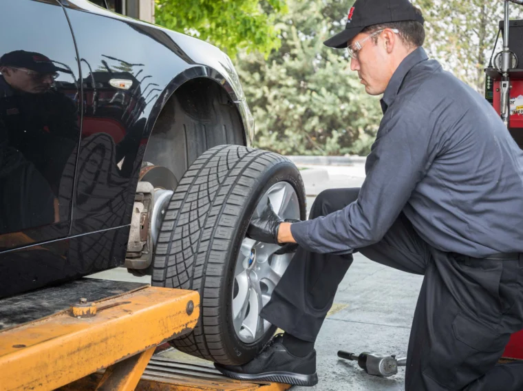 A Grease Monkey mechanic in uniform and gloves installs a tire on a car lifted by a hydraulic platform in an auto repair shop offering State Inspection Services.