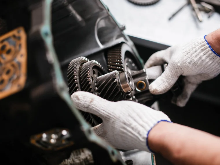 A person wearing white work gloves adjusts gears inside a mechanical assembly—likely part of an engine or transmission services setup.