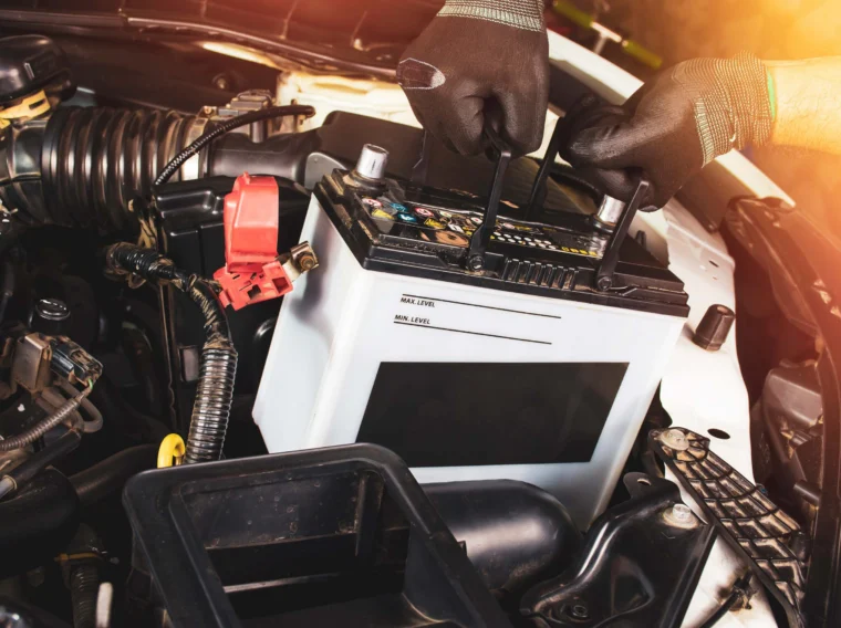 Two hands holding a car battery underneath the hood of a vehicle.